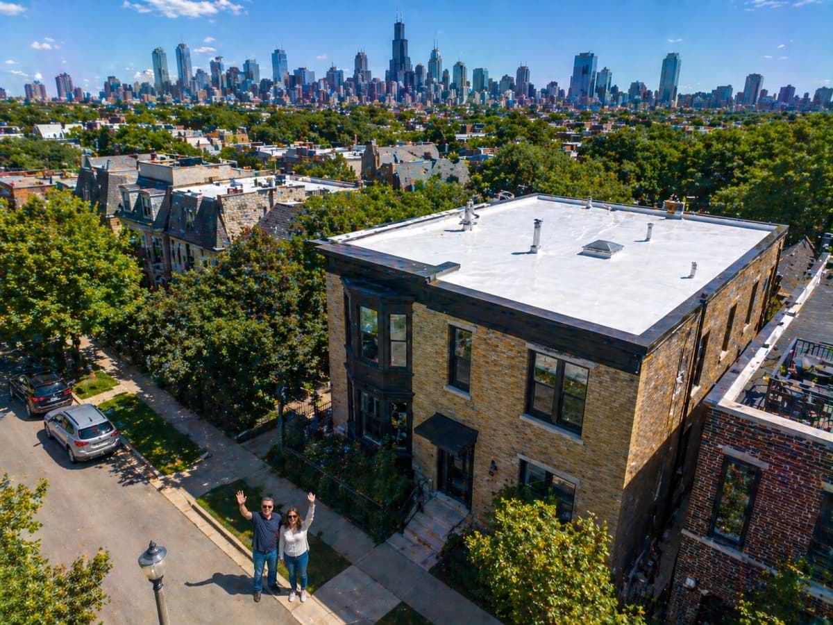Aerial view of a Chicago two-flat with a newly installed white TPO flat roof, with the downtown Chicago skyline in the background. Homeowners stand on the sidewalk after a complete 2026 roof replacement project.